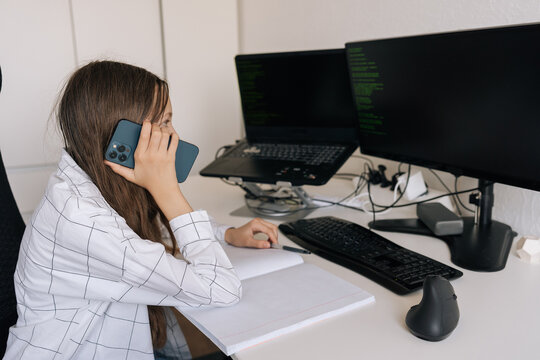 Side view of young girl working remotely from homeworking, communicating on smartphone while writing in notebook near laptop and monitor displaying programming code.