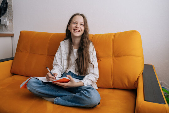Happy young girl sitting cross-legged on orange sofa, writing in notebook. She looking directly at camera with joyful smile, suggesting positive experience with remote learning creative journaling.