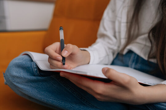 Unrecognizable person writes notes in planner or diary. Closeup on hand holding pen over paper. Student studies, doing homework or journaling. Calm, focused mood in comfortable home setting.