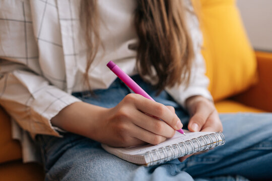 Close-up of young person sitting comfortably, holding vibrant pink pen and writing down thoughts in spiral notebook, focusing on creativity, learning, planning.
