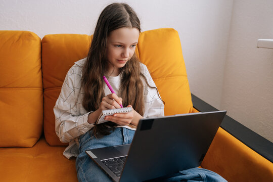 Young girl focusing on laptop screen while taking notes in small spiral notebook, engaged in distance learning or online education from comfort of comfortable couch at home.