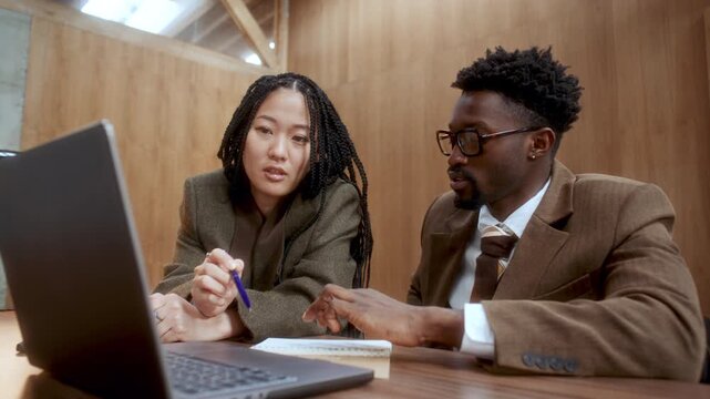 Young Asian woman and her African American business partner working on laptop and discussing notes at desk in office with wooden walls