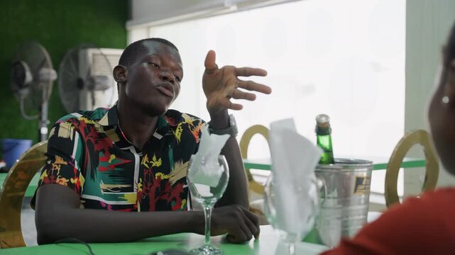 Black man on date at table, shy playful gestures and candid conversation with partner across green tablecloth ice bucket, wine bottle, glassware and folded napkin create intimate casual bistro scene