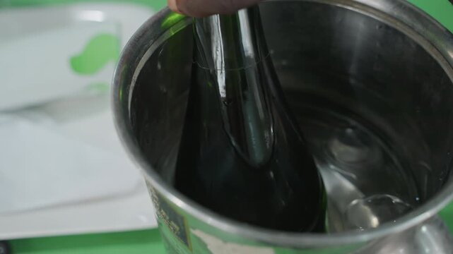 Closeup black bottle chilling in bucket, stainless container half filled with water and ice, dark bottle neck rising with condensation, reflective surface catches green background, intimate