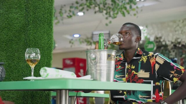 Black man sipping wine at table enjoys slow sip from goblet beside ice bucket and bottle in green themed indoor bistro, relaxed smile suggesting successful date or quiet celebration