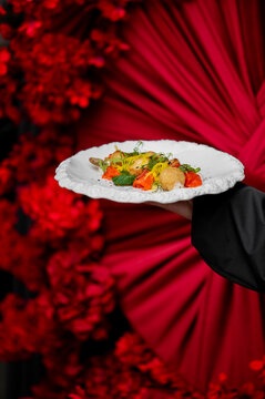 A professional waiter in a black suit holds a white textured plate featuring a gourmet seafood appetizer against a vibrant, out-of-focus background of red flowers and draped fabric.