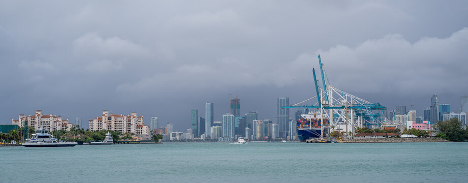 Port of Miami and Fisher Island under cloudy sky. Wide view of Biscayne Bay with gantry cranes at the port and residential buildings on Fisher Island.