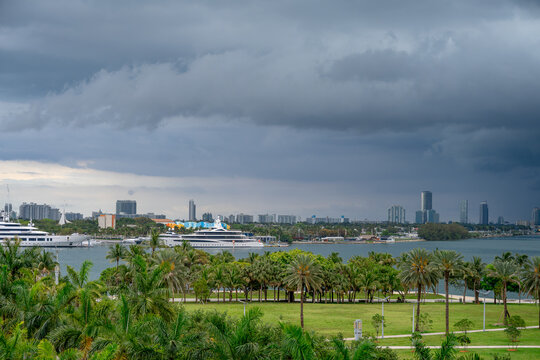 Luxury yachts in Miami harbor under stormy grey sky. Waterfront view of large yachts and cruise ships in Biscayne Bay with dark clouds and tropical park landscape.