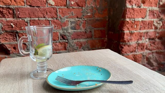 Empty turquoise plate with fork, glass tumbler holding lemon slice on linen tablecloth against exposed brick, intimate cafe vibe
