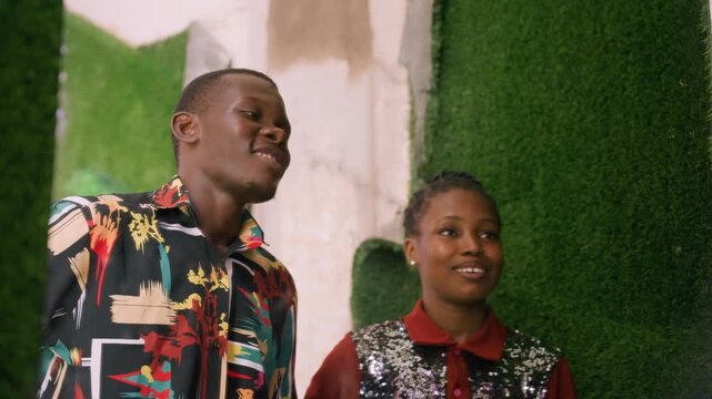Playful african couple sharing flirtatious moment by green wall, man leans for kiss, woman laughs and covers mouth, matching watches, patterned shirt, candid dating vibe, joyful romantic energy