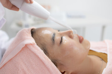 Naklejka premium Soft light glows on calm face. Gentle device delivers soothing mist. Eyes closed, expression serene. Pink towel wraps tenderly around head. Perfect for wellness, spa, or beauty branding 