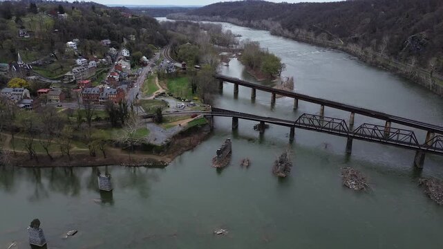 Lower Town Harpers Ferry sits between Potomac Shenandoah Rivers beneath Maryland Heights rail crossing. Brick blocks, metal roofs, soft spring canopies, river sheen define textures in WV