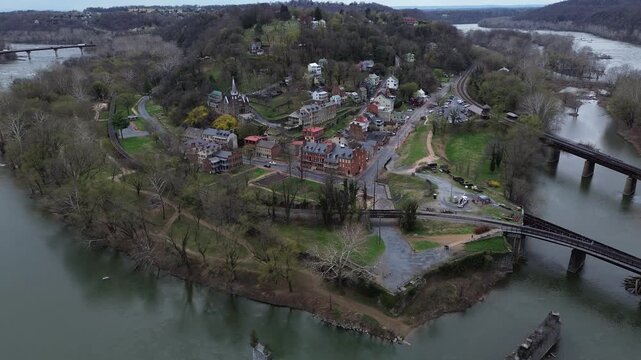 Harpers Ferry riverfront curves along Potomac beside rail bridge narrow historic streets below wooded ridge. Mixed roofing, early leaf-out, warm hillside tones shape textures in WV