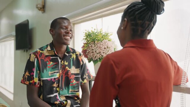 Black couple sharing shy smiles by window, patterned shirt man and red blouse woman exchange playful banter, potted plant, sunlit interior, cozy conversation, gentle laughter, romantic tension