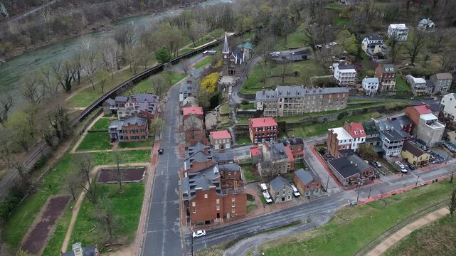 Harpers Ferry hillside blocks step down toward river rail line with tightly clustered brick stone structures. Weathered roofs, muted spring trees, angled valley light define textures in WV