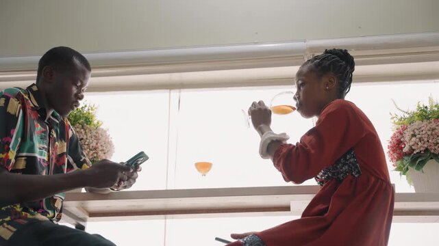 Black couple intimate window date scene, sharing orange cocktail by sunlit counter, floral bouquets on sill, man in patterned shirt scrolling phone then engaging, woman in red dress sipping, soft