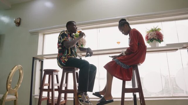 Black couple playful barstool date scene, clinking cocktail glasses across sunlit counter, man in patterned hoodie offering toast, woman in red dress laughing, midcentury cafe interior, tall stools,
