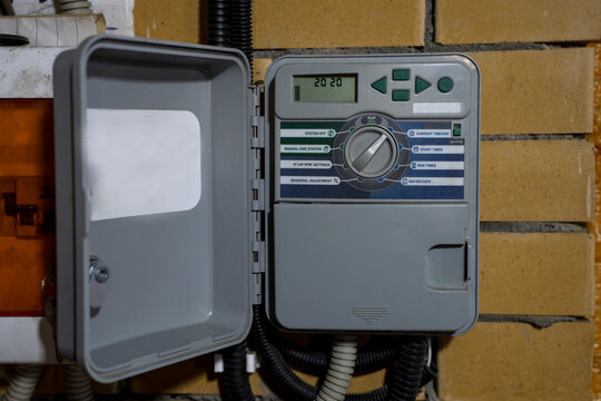 Person adjusting irrigation controller for automatic watering system, managing sprinkler zones via digital control panel mounted on brick wall.