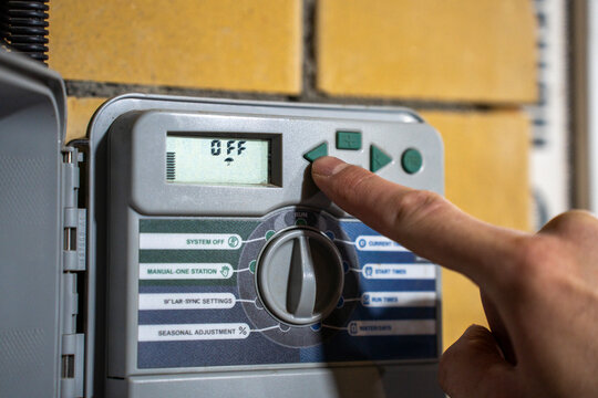 Finger pressing button on irrigation controller panel, adjusting automatic watering system settings with digital display and selector dial.