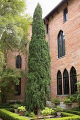 Fototapeta premium Tall green tree stands beside brick building with pointed windows in warm daylight while garden plants surround the base