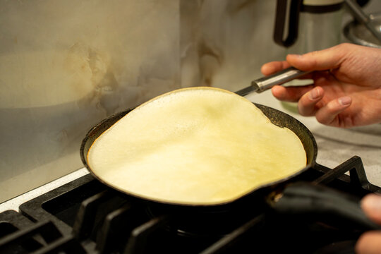Thin pancake cooking in frying pan on gas stove, close-up of homemade crepe preparation in kitchen with hand holding pan handle.