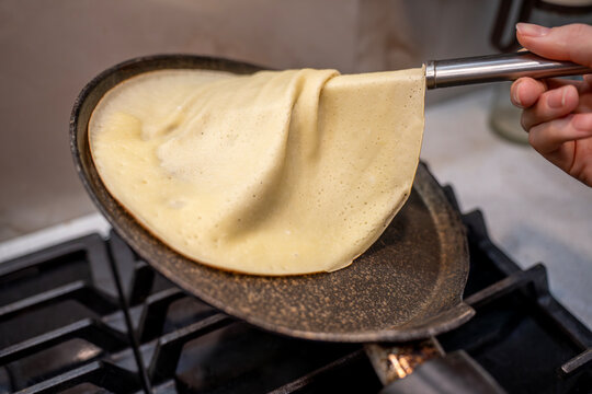 Hand flipping thin pancake with spatula in frying pan on stovetop, close-up of homemade crepe cooking process in kitchen.