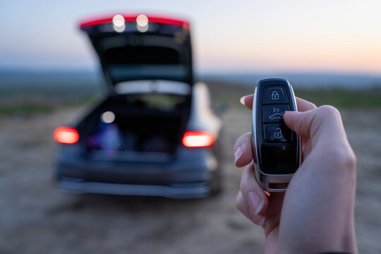 Hand holding car key fob near vehicle with open trunk, blurred rear lights and evening outdoor background.