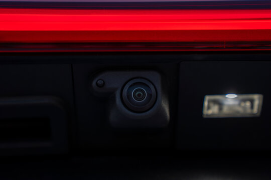 Close-up of rear view camera mounted on the tailgate of a modern car, positioned near the license plate light under red taillight strip.