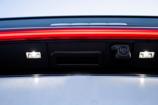 Close-up of rear view camera and license plate lights on a modern car tailgate, positioned below a red taillight strip on the rear body.