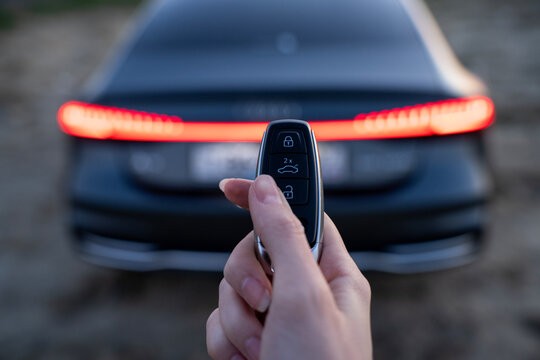 Hand pressing trunk button on car key fob near rear of vehicle, with illuminated tail light and blurred automobile background outdoors.
