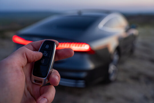 Hand pressing button on car key fob near blurred vehicle with illuminated rear lights outdoors at dusk.