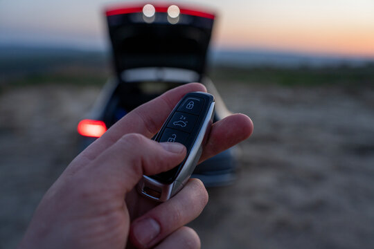 Finger pressing button on car key fob in front of vehicle with open trunk, close-up of remote control use at dusk.