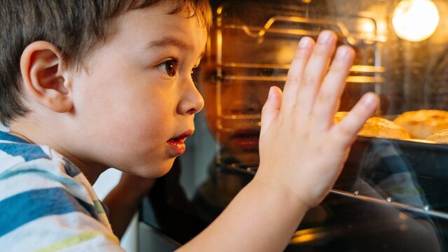 A little boy with bright eyes and an eager expression presses his hand against the oven glass, captivated by the golden pastries baking inside. It's a moment of wonder and anticipation.