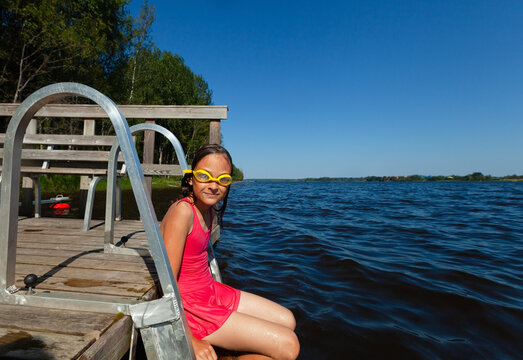 Girl perches on a metal ladder at a dock with legs dangling in water on a sunny summer day. Child in red swimsuit and yellow goggles relaxing by the lake. Kid enjoys carefree outdoor leisure and swim