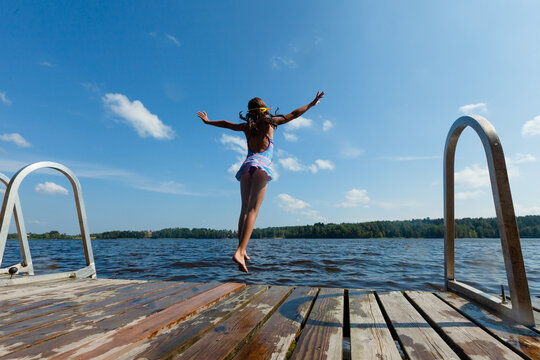 Child leaps off wooden dock into lake continuing beloved family vacation tradition at lake house. Little girl enjoys summer rituals and lakeside fun on day with loved ones nearby. Kid creates lasting.