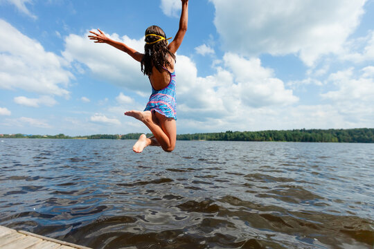 Girl leaps off a dock mid-air on a sunny summer day. Child jump from pier in a joyful dynamic pose above the water. Kid experiences carefree freedom and refreshing outdoor recreation