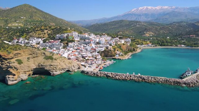 Breathtaking Agia Galini Crete. Aerial 4K Drone reveal from white village and turquoise port to hidden sea caves in yellow sandstone cliff. Snow-covered mountains background. Greece spring travel