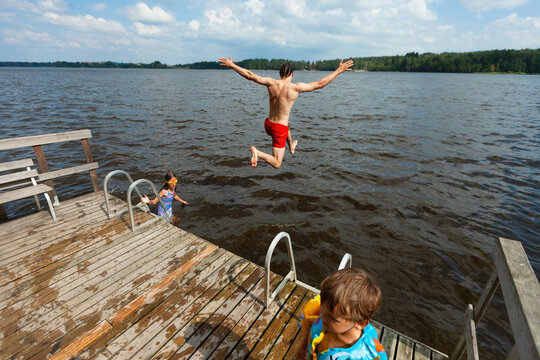 Father with children at the lake enjoying family summer vacation on sunny day. Man in red trunks and kids gather by the water and dock outdoors. Happy family shares joyful active recreation holidays