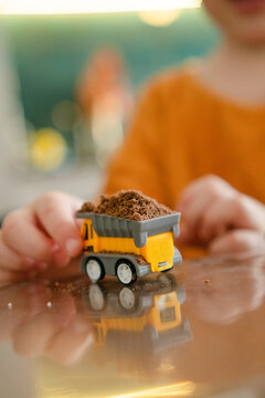 Playful toddler child holds yellow toy dump truck with dirt in small hands. Kid explores texture through sensory tactile play on reflective indoor surface. Curious youngster enjoys practical learning