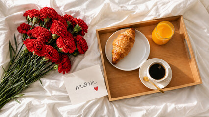 A top-view cinematic composition of a Mother's Day surprise: a wooden tray with a croissant, coffee, and juice next to red carnations and a "Mom" card on soft white bedding in bright morning light.