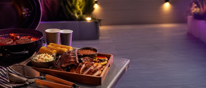Outdoor barbecue feast with grilled ribs, sausages, corn, coleslaw, sauce and drinks beside a glowing grill on a night patio with copy space