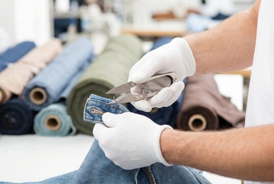 Garment worker wearing white gloves using thread snips to trim loose threads on blue denim jeans
