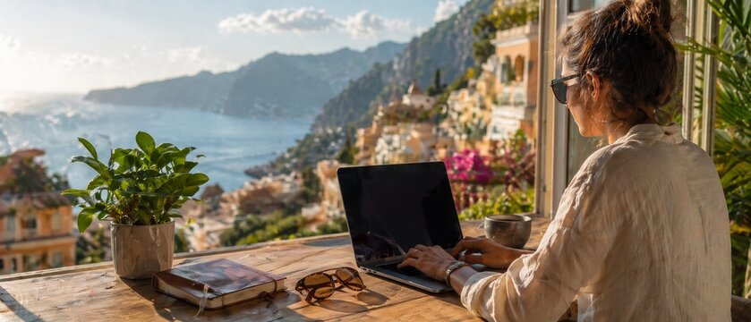 Woman using laptop at sunny window desk overlooking coastal town, sea and mountains with potted plant, notebook, sunglasses and warm travel light