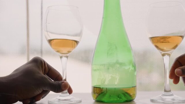 Romantic wine date with black couple sharing amber wine from green bottle and stem glasses by sheer window hands reach to clink glasses over sunlit table, condensation on bottle, intimate cozy mood,