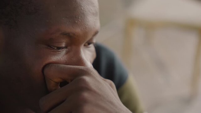 Black man thoughtful close portrait caf, shy date emotion with hand near mouth, glances upward and slight smile, tear shimmering on lower lid, striped sweater and wooden chair blur, warm natural light