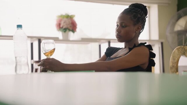 Black woman at table holding wine in soft light, braided hair and elegant dress, water bottle and floral vase on windowsill quiet postdate reflection with thoughtful gaze, close framing and cinematic