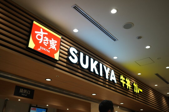 Bright sign glows above a modern restaurant interior. Wood paneling adds warmth to the ceiling. Customers linger near the entrance. Lighting highlights the sign's bold lettering