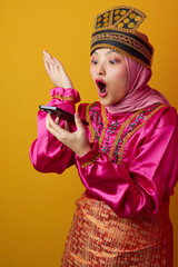 Naklejka premium Surprised Young Indonesian Asian Woman in Bright Traditional Costume and Hijab Reacting While Holding Phone