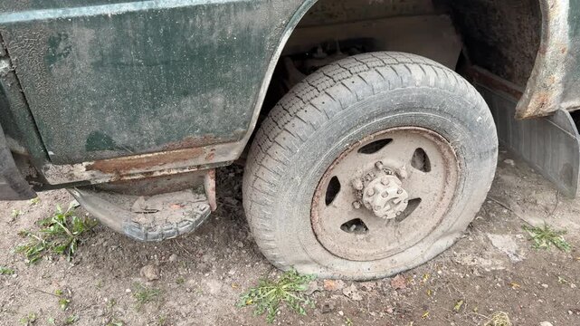 Abandoned dark green minibus with punctured tire, close-up of muddy wheel and spare rim tucked under chassis, heavy rust patches