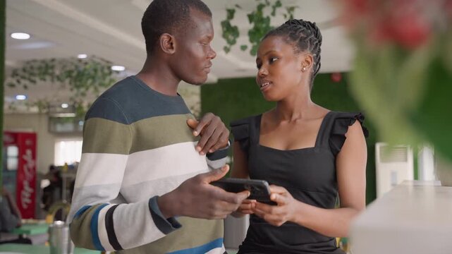 Black couple sharing phone and laughing at cozy cafe table, man points at screen while woman smiles, plants and soft lights frame intimate playful moment, casual outfits and lighthearted flirtation